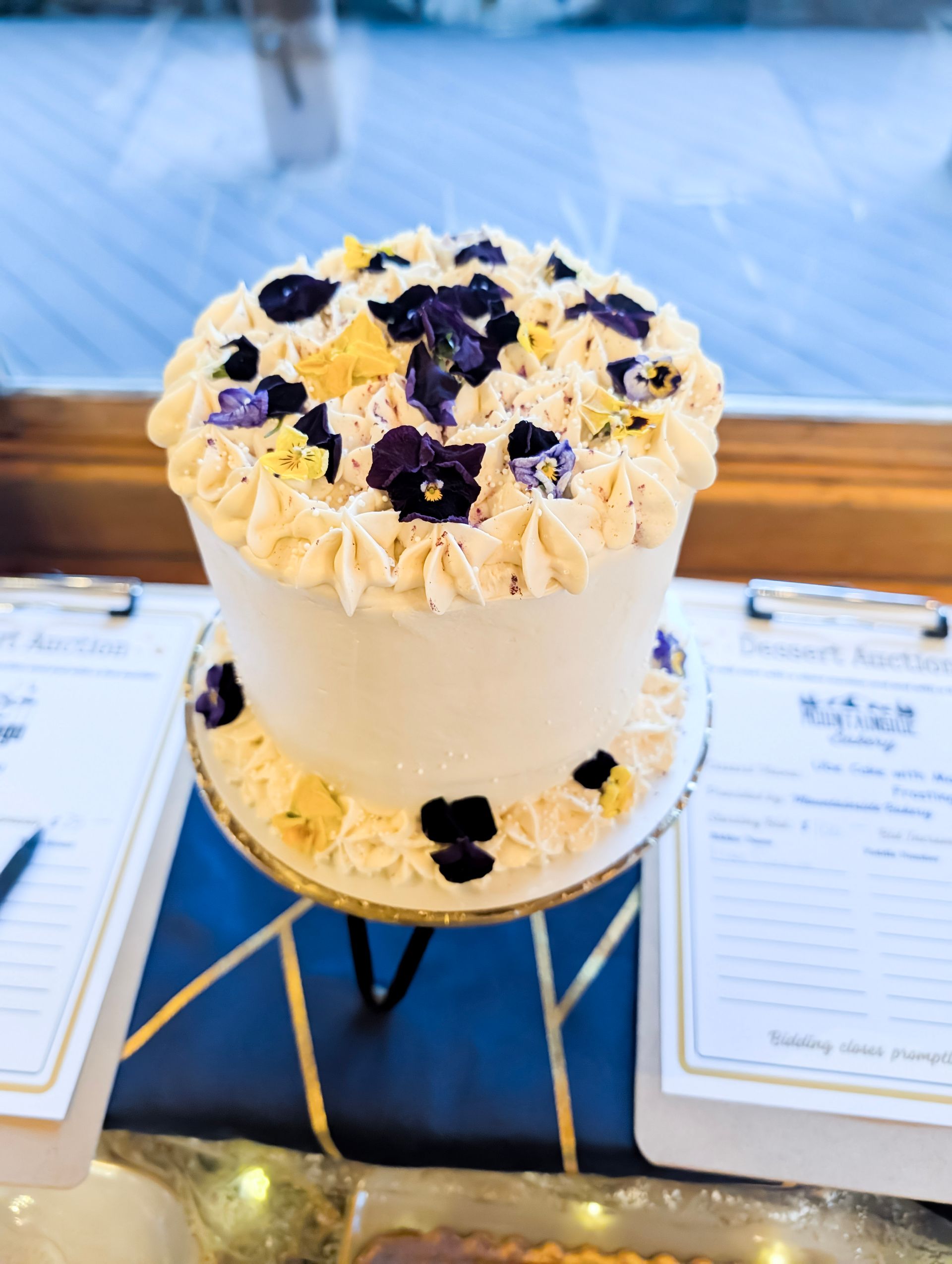 White frosted cake topped with dark berries and flowers on a stand, displayed beside menus on a table