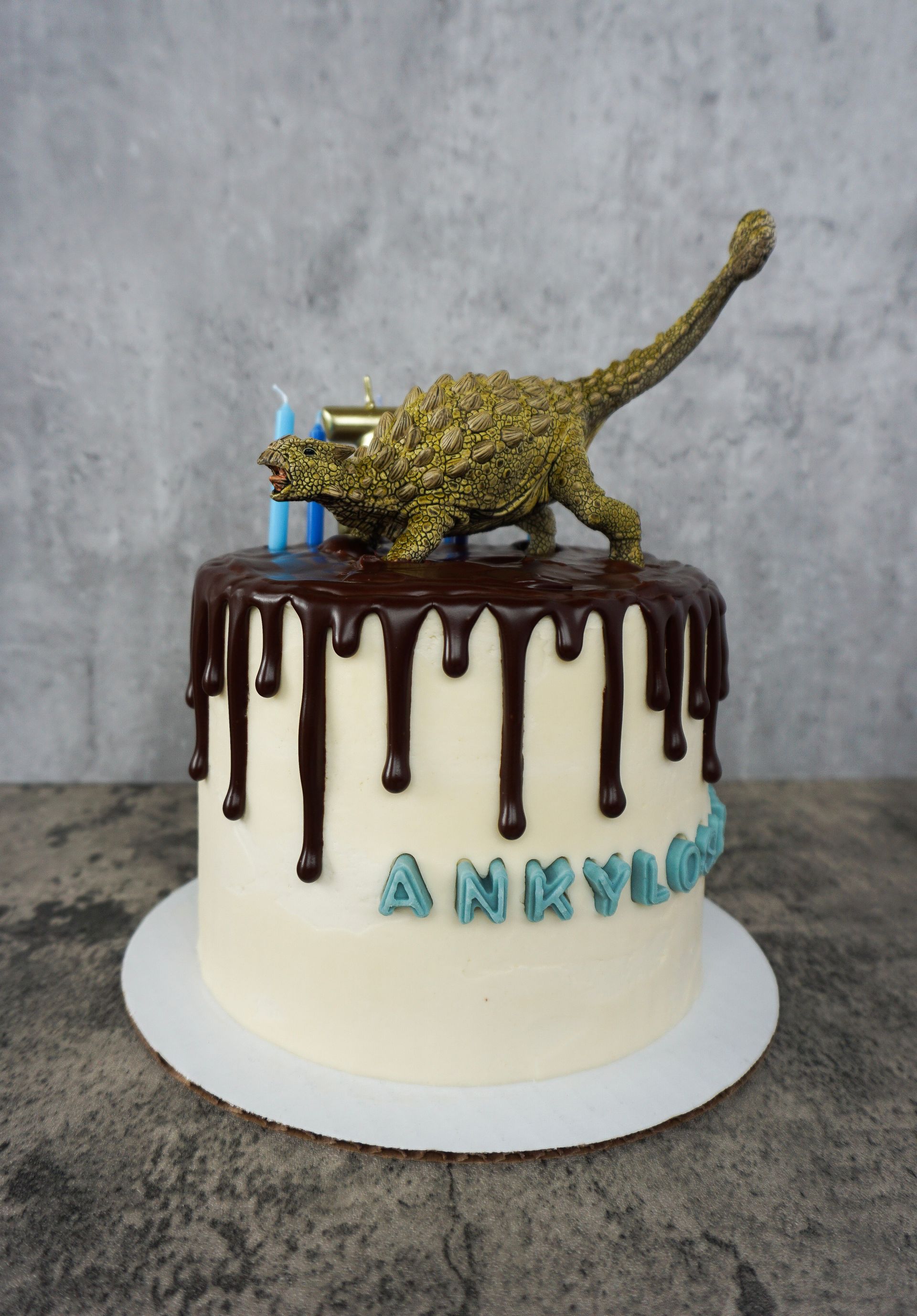 Cake topped with a gold dinosaur and dark chocolate drips, with blue “ANNV” letters on the front