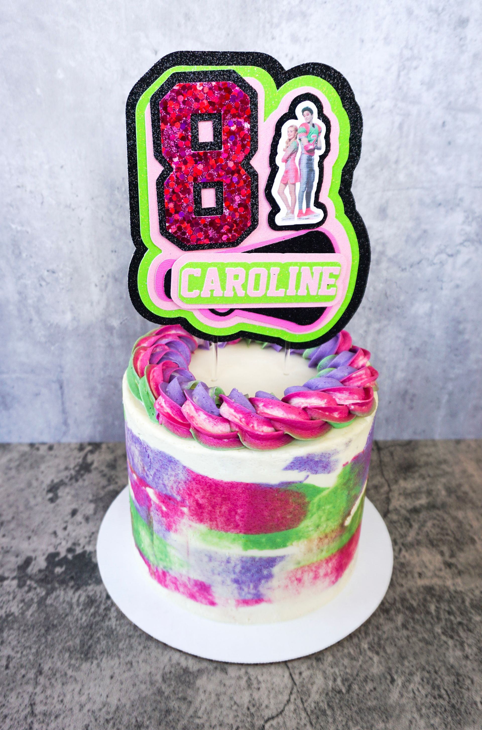 Bright birthday cake with pink, green, and white frosting, topped with “8” and “CAROLINE” decorations