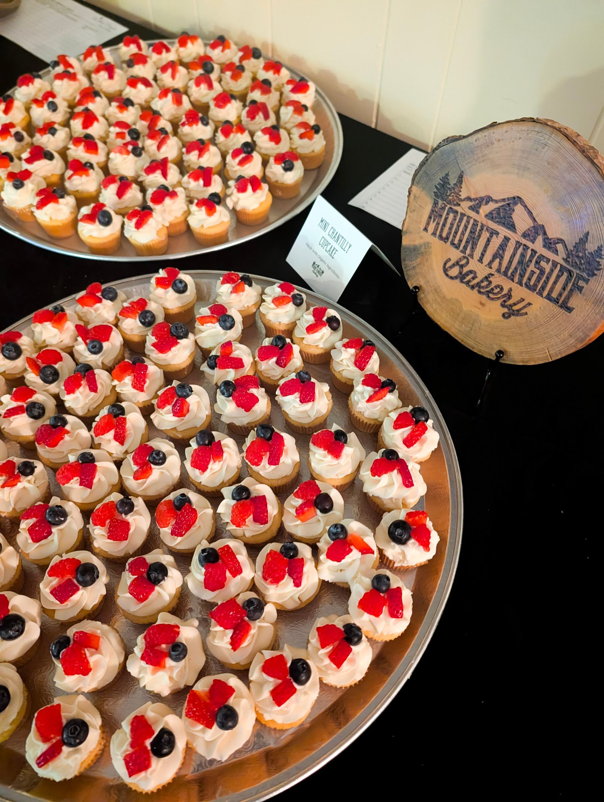 Trays of red-topped cupcakes on a table beside a rustic wooden sign at a dessert display