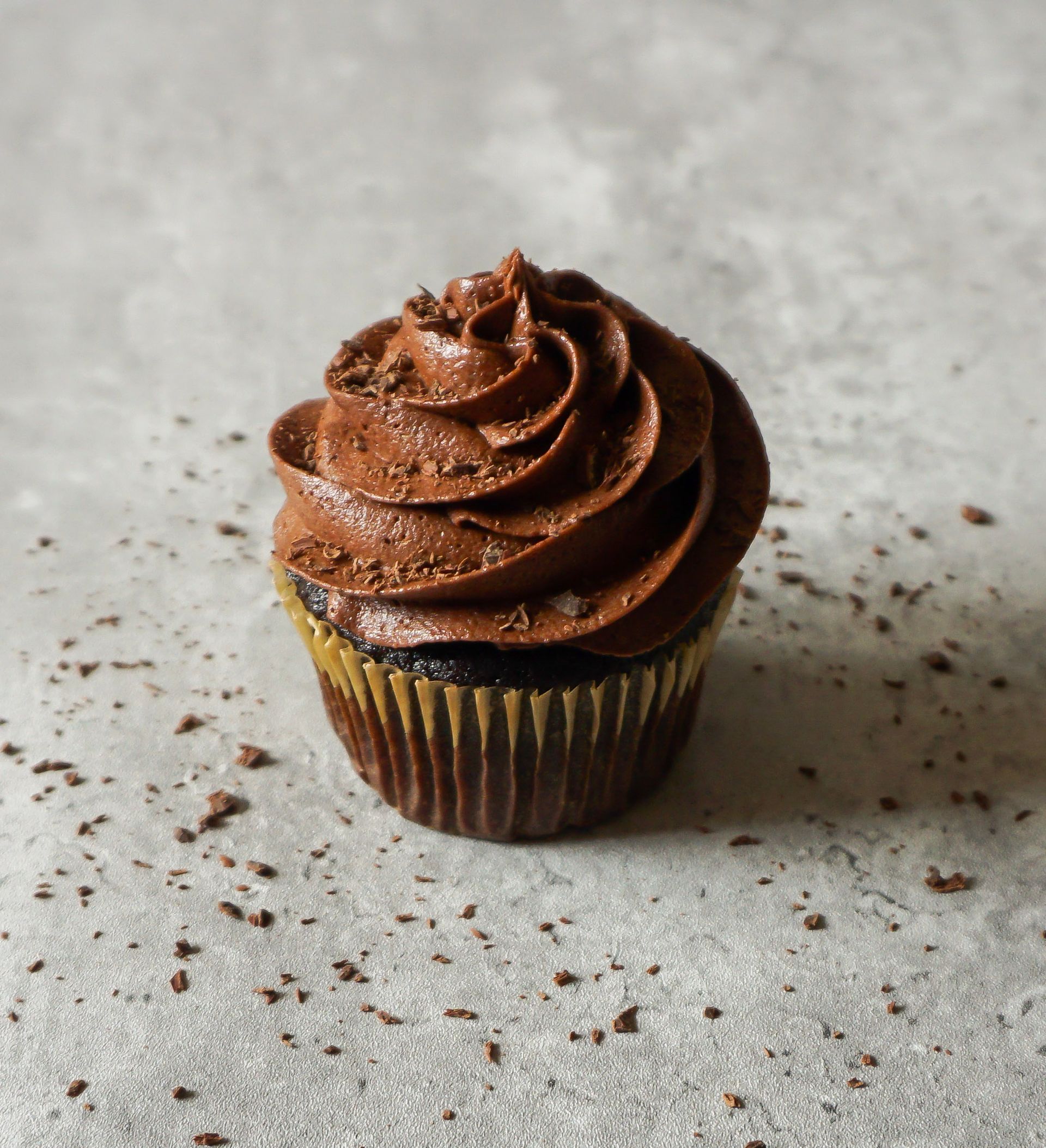 Chocolate frosted cupcake in a striped wrapper on a gray surface, with scattered cocoa crumbs.