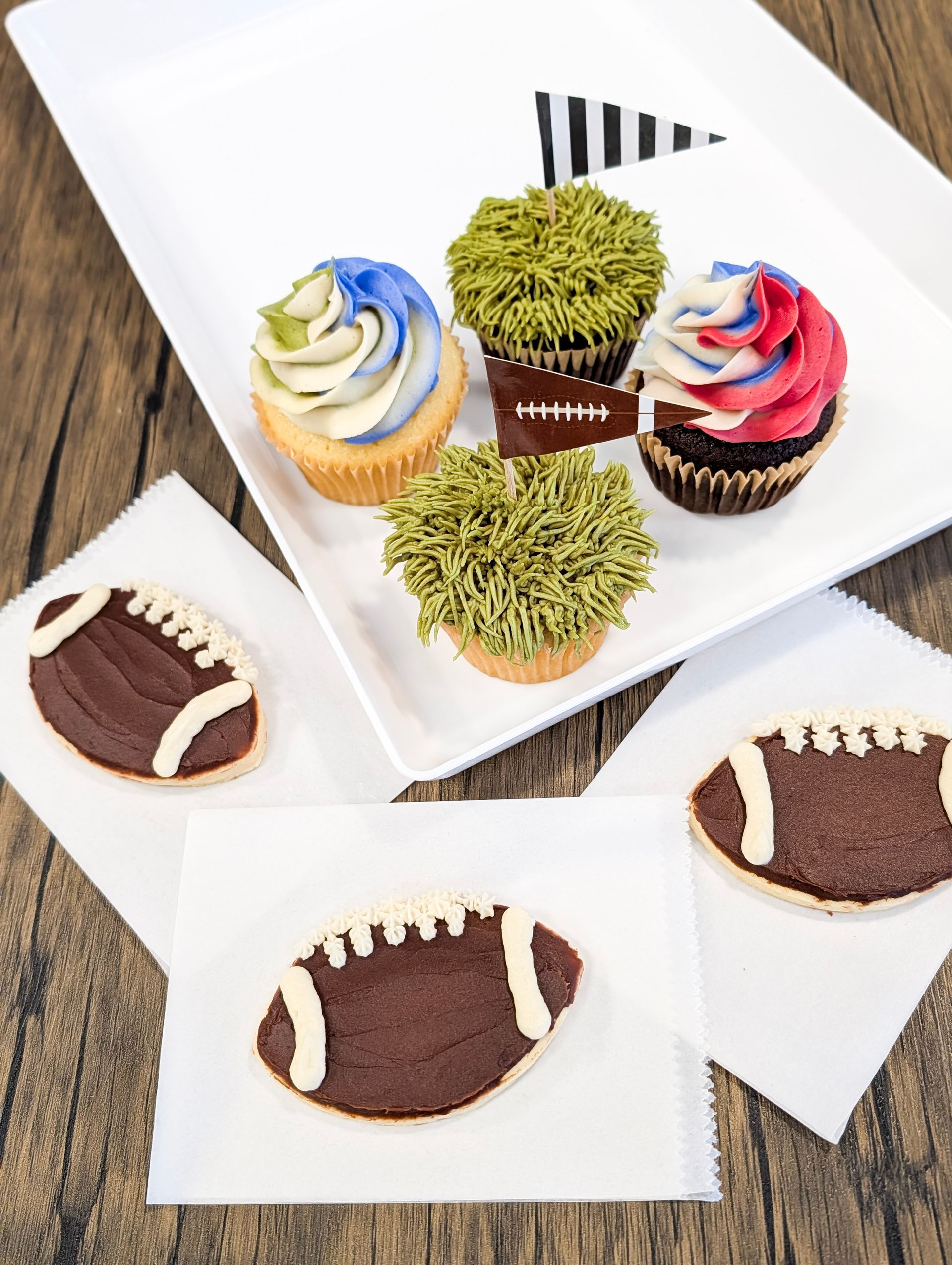 Assorted decorated cupcakes on white trays, including football-shaped chocolate cakes and colorful frosted cupcakes