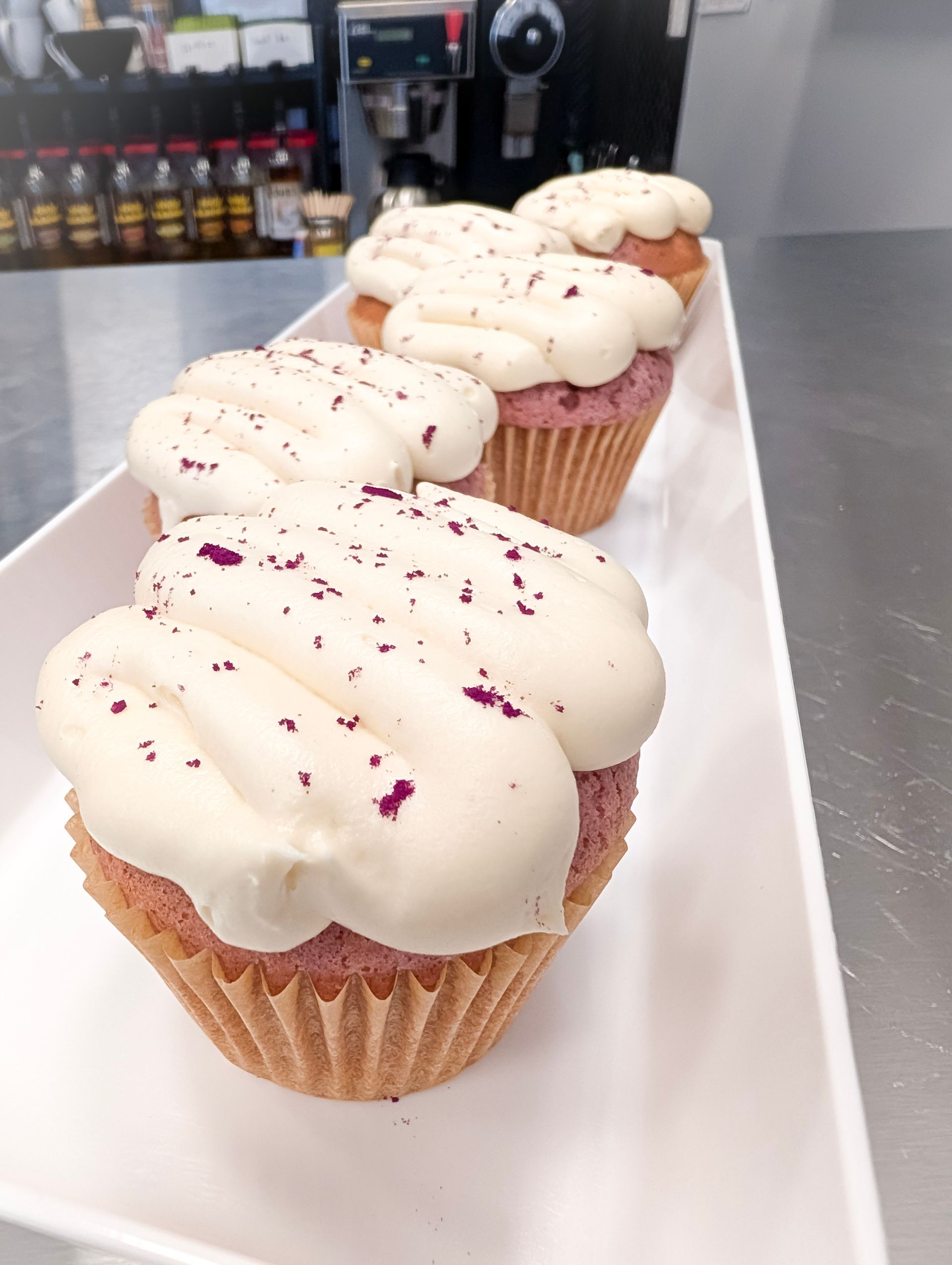 White-frosted cupcakes with pink sprinkles lined on a tray in a bakery display.