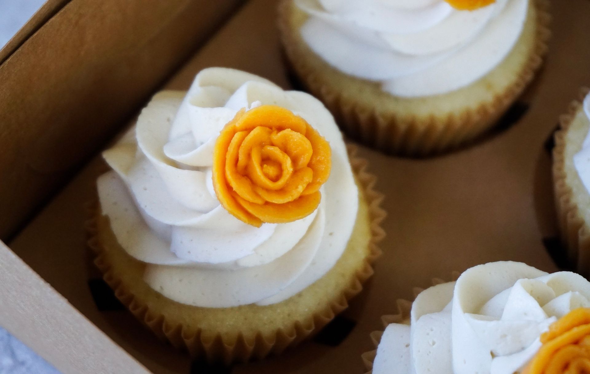 White cupcakes with swirled frosting and orange flower toppers in a cardboard box