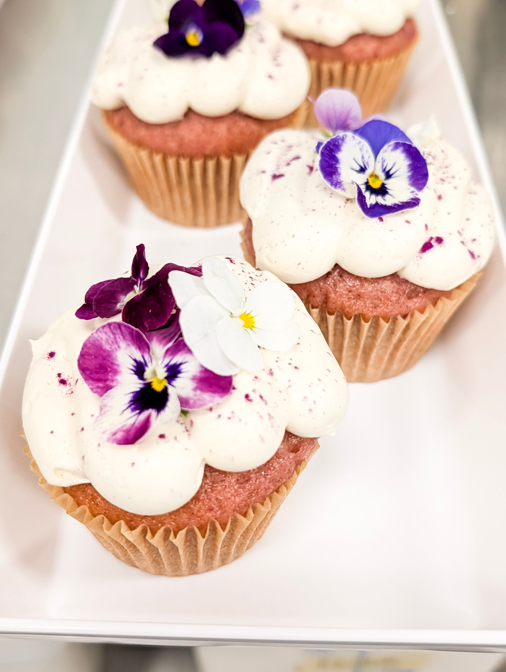 Three frosted cupcakes topped with purple and white edible flowers on a white tray