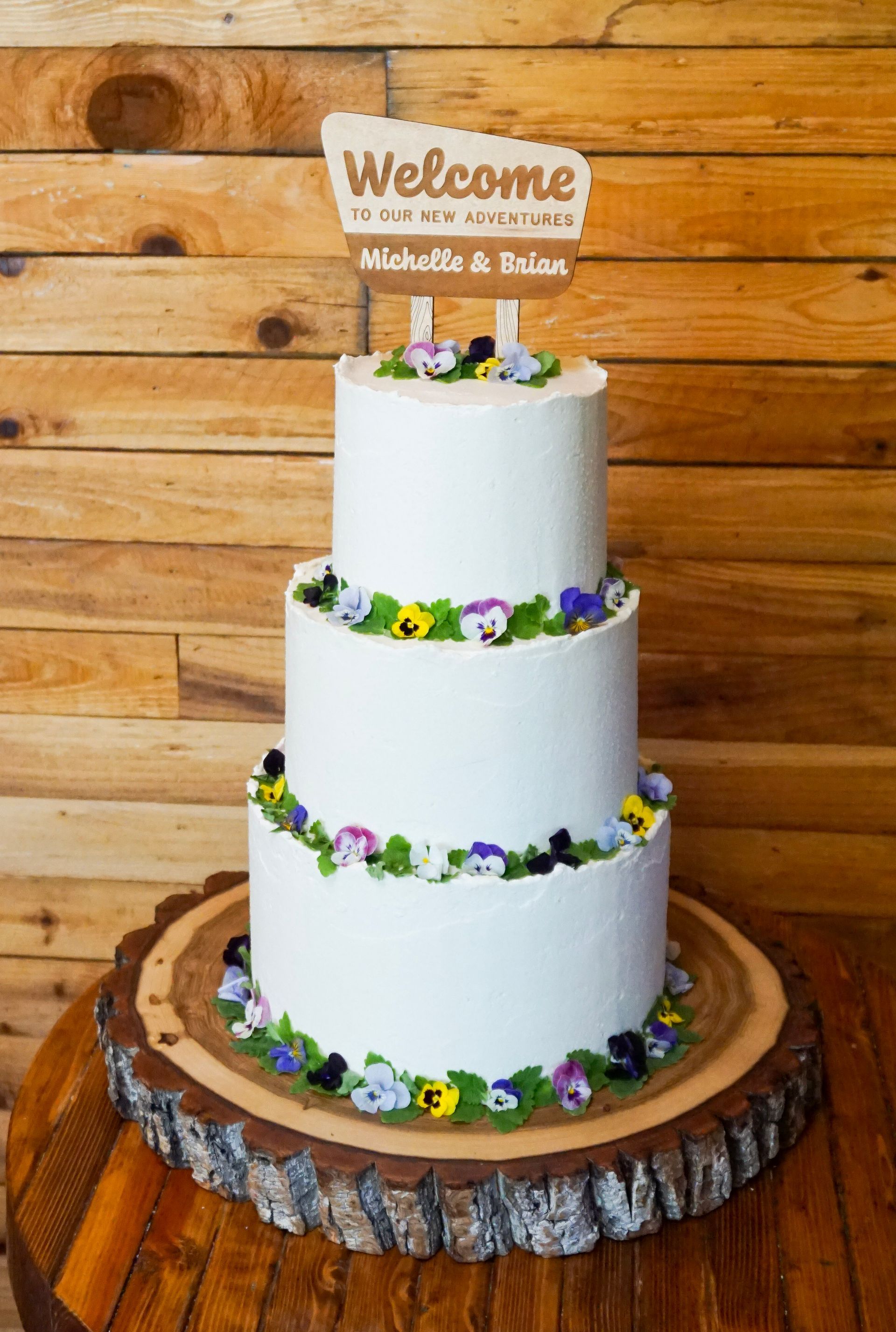 Three-tier white wedding cake with purple and yellow flowers and a “Welcome” topper on a wooden barrel stand