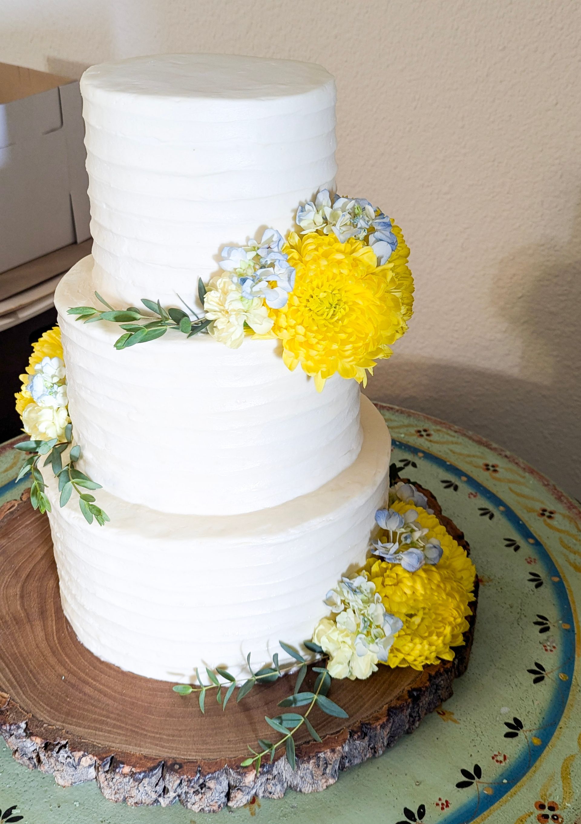 Three-tier white cake with yellow and white flowers on a decorated round cake board