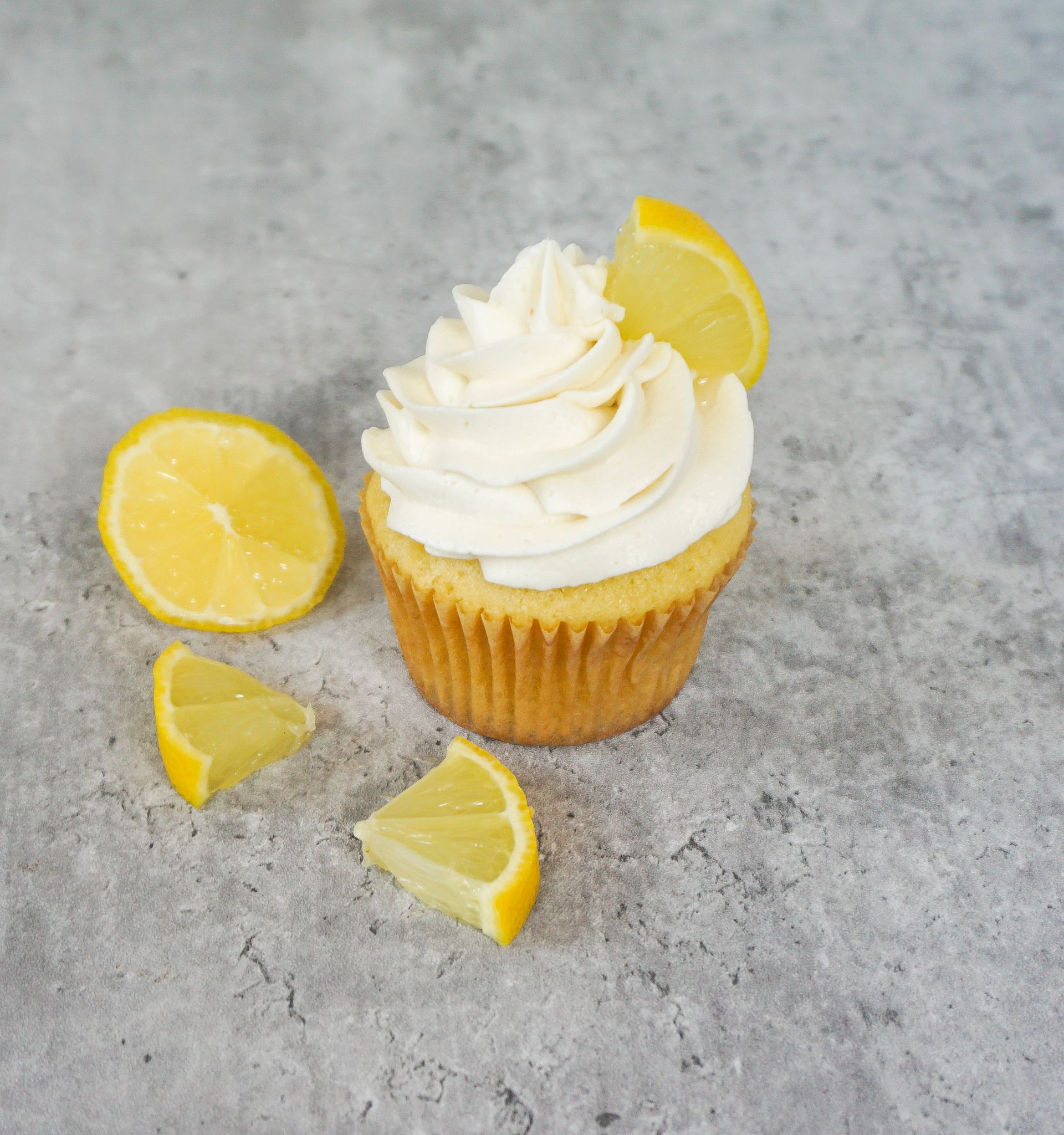Lemon cupcake topped with white frosting, surrounded by lemon slices on a gray surface