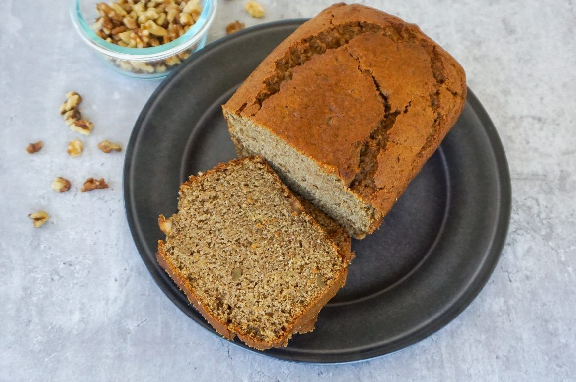 Sliced loaf of banana bread on a dark plate with chopped walnuts nearby