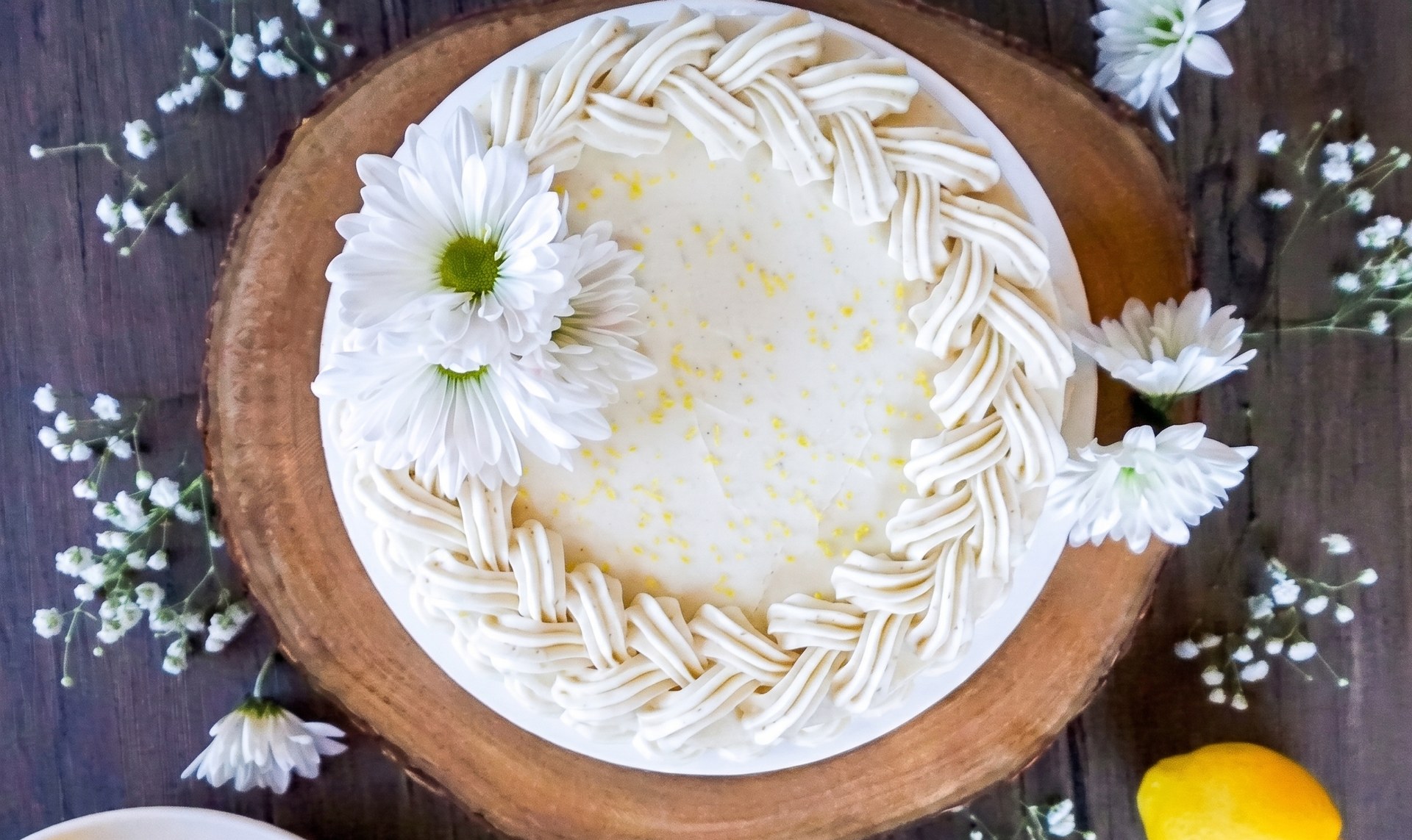 White frosted cake with daisies on a wooden board, surrounded by small flowers on a dark table.