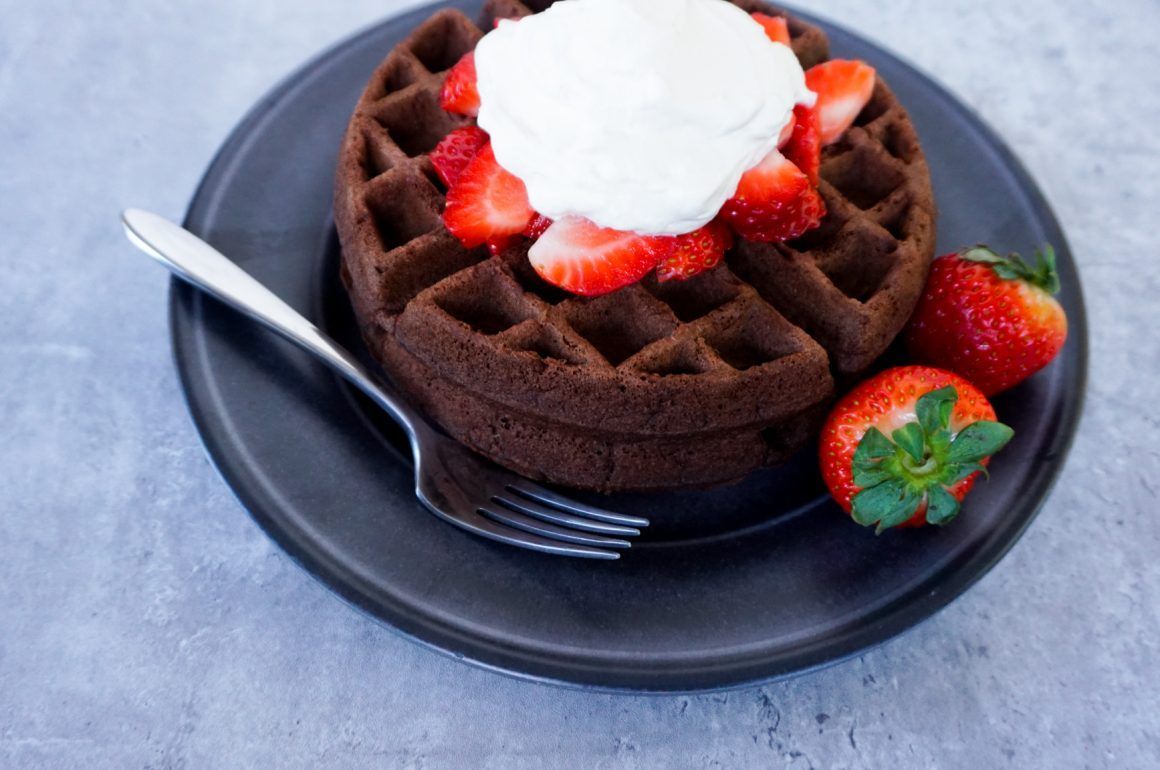 Chocolate waffle topped with whipped cream and strawberries on a black plate with a fork