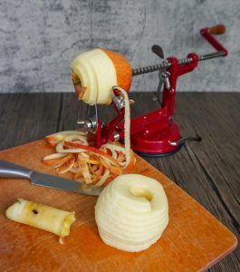 Red apple being peeled with a hand-crank peeler on a wooden cutting board, peel curls below.