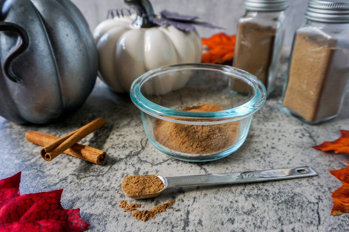 Glass bowl of brown spice on a stone table with cinnamon sticks, a spoon, and jars in the background