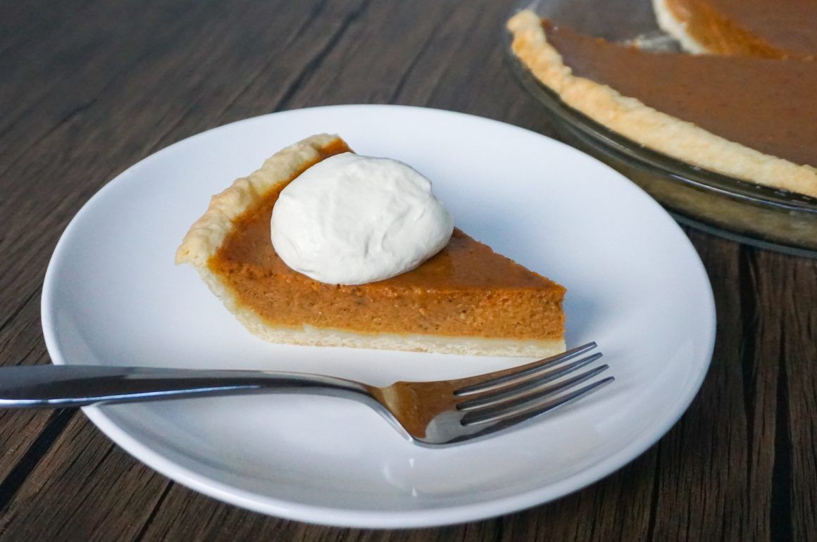 Slice of pumpkin pie with whipped cream on a white plate, with a fork on a dark wooden table