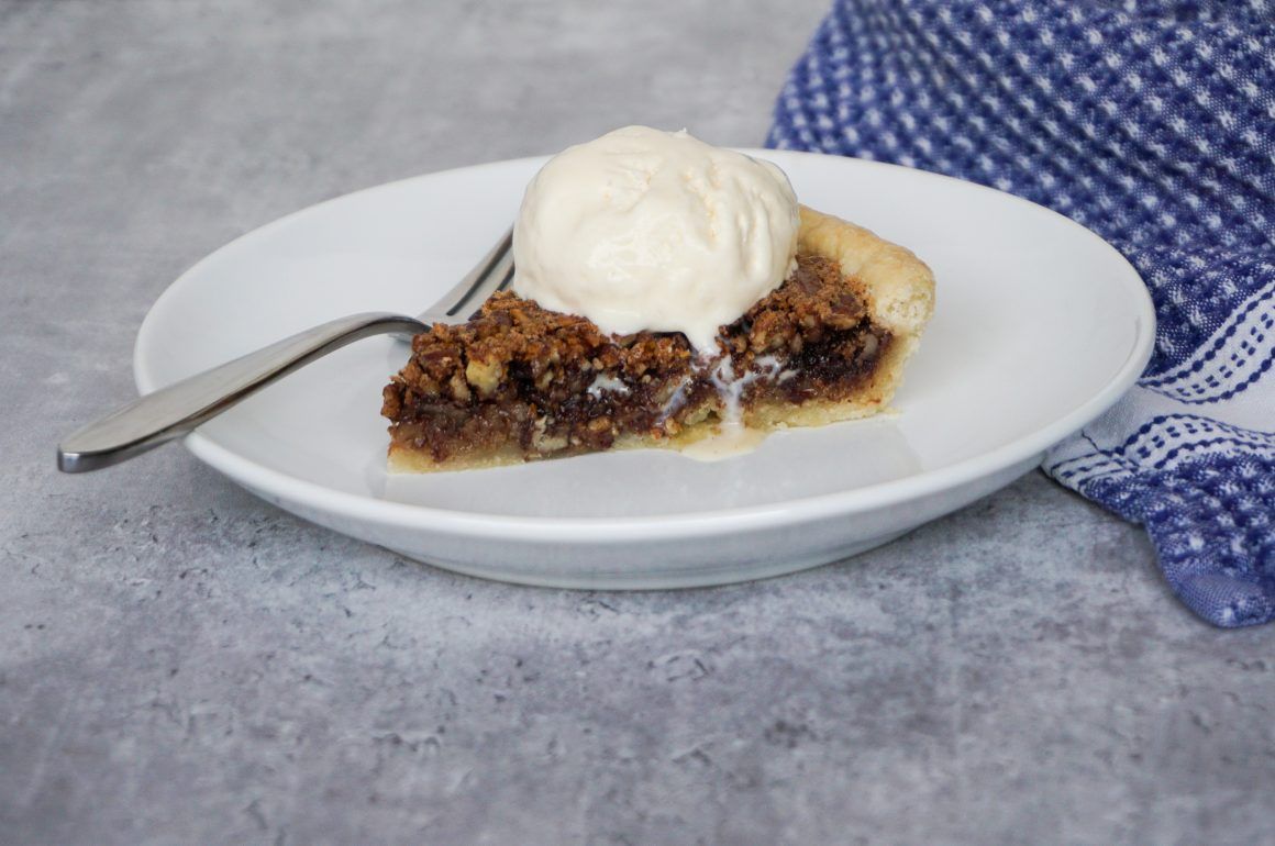 Slice of pie with vanilla ice cream on a white plate, beside a fork and blue cloth napkin
