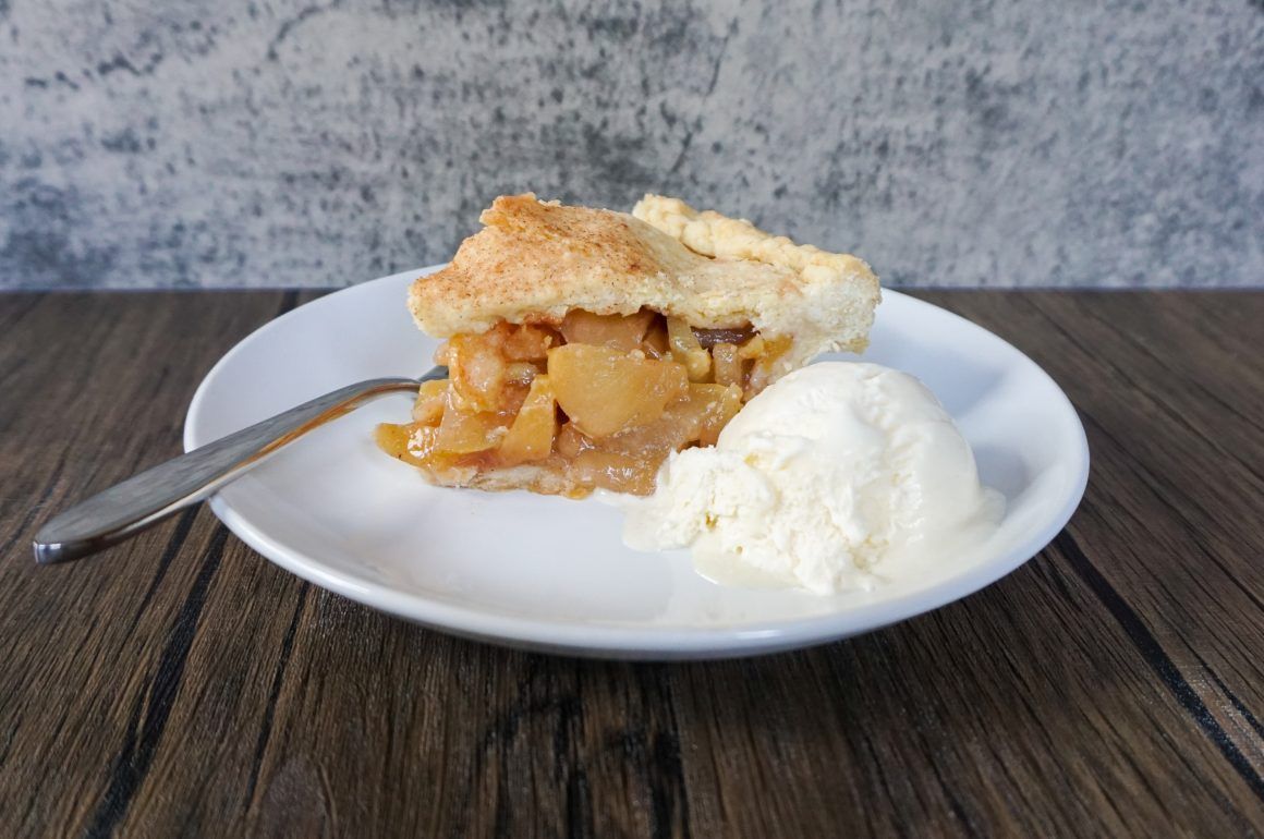 Slice of apple pie on a white plate with a scoop of vanilla ice cream and a fork on a wooden table