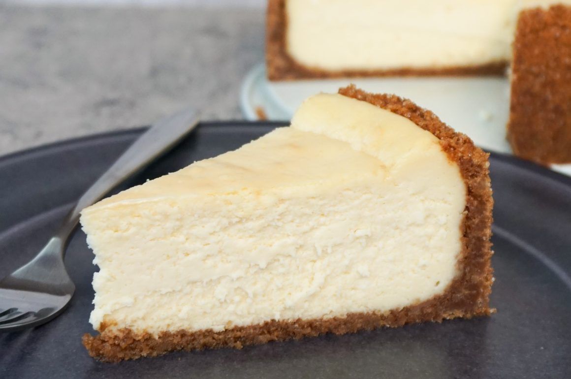 Slice of cheesecake on a dark plate with a fork, with more cake in the background