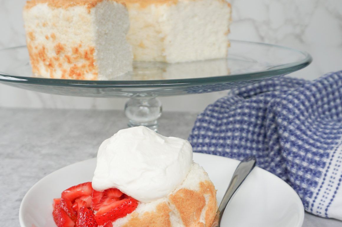 Slice of strawberry shortcake with whipped cream on a white plate, with a whole cake in the background
