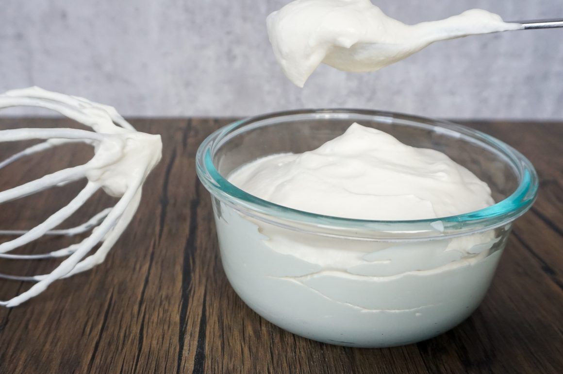 Glass bowl of white whipped cream with a whisk beside it on a wooden surface