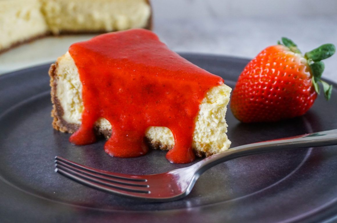 Slice of cheesecake topped with strawberry sauce on a plate, with a fork and fresh strawberry beside it