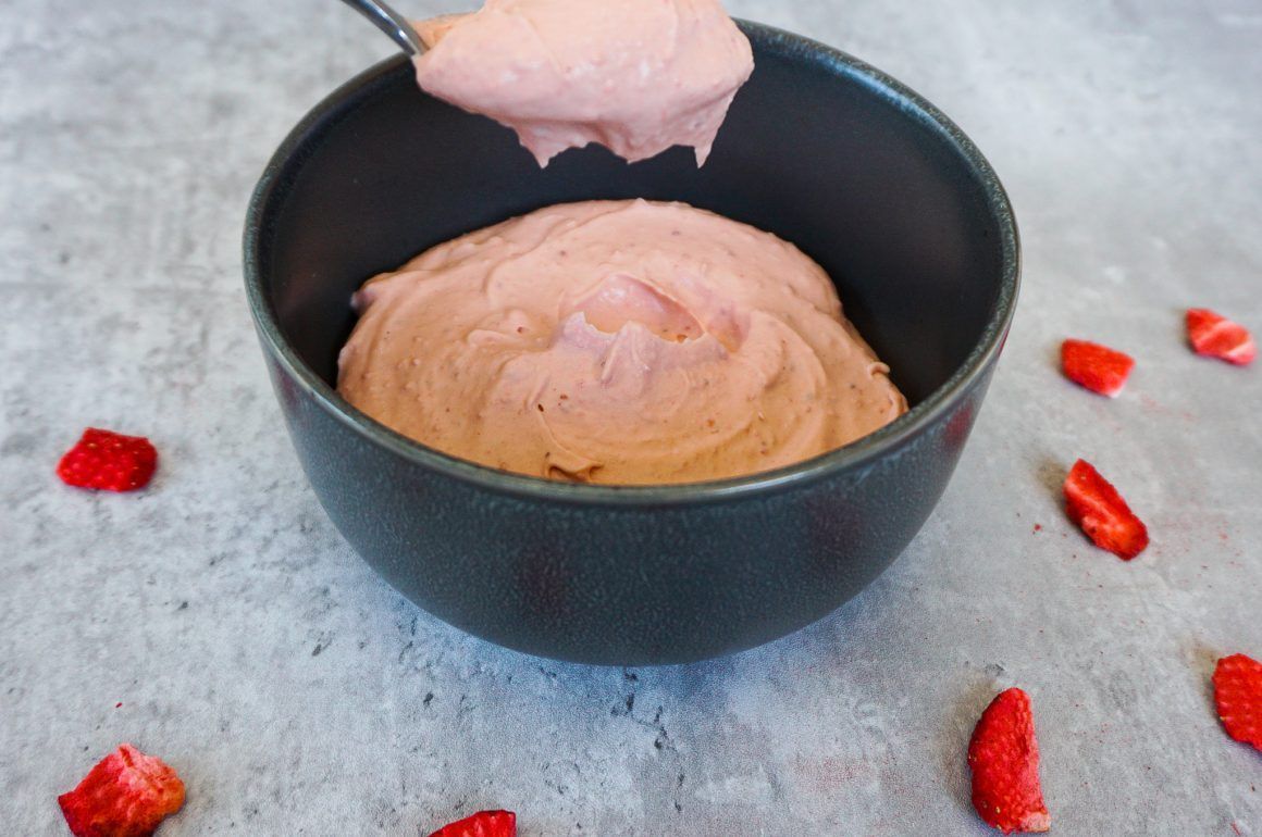 Scoop of pink ice cream in a black bowl, with strawberry pieces on a gray surface