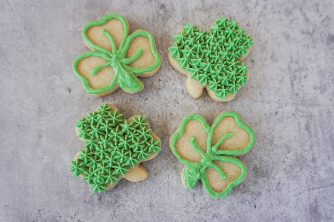 Four green shamrock-shaped cookies on a gray background, two decorated with piped leaves and two with star-like frosting.