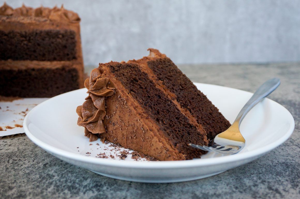 Slice of chocolate layer cake with frosting on a white plate, with a fork beside it