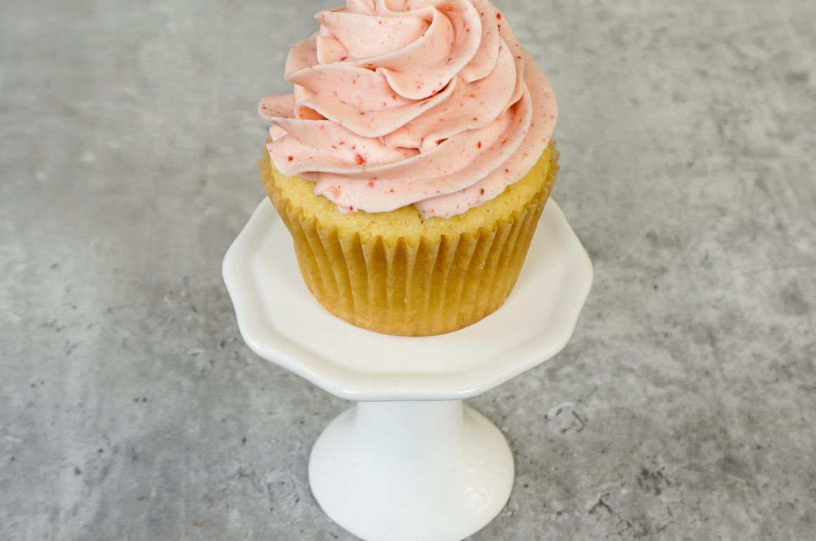 Vanilla cupcake with pink frosting on a white pedestal against a gray background
