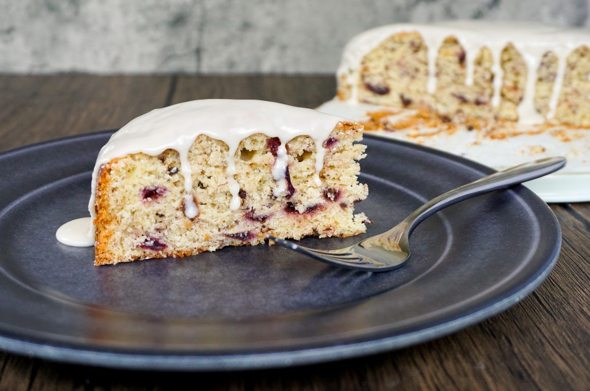 Slice of crumb cake with white icing on a dark plate, with a fork and a larger cake in the background