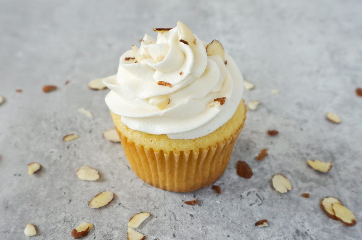 Vanilla cupcake with white frosting on a gray surface, surrounded by sliced almonds.