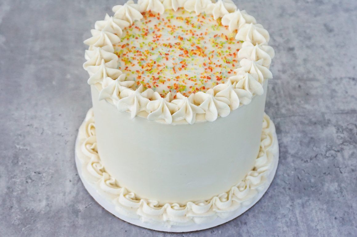 White frosted cake with piped cream rosettes and colorful sprinkles on top, on a white cake board