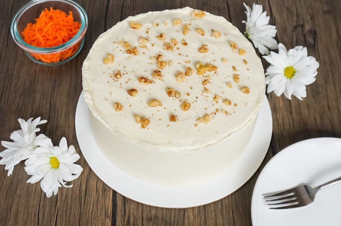White frosted cake with nuts on a plate, garnished with flowers and a bowl of orange shreds nearby