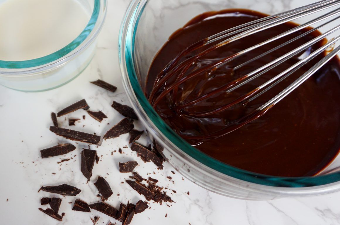 Whisk in a bowl of melted chocolate with chocolate shavings on a white surface