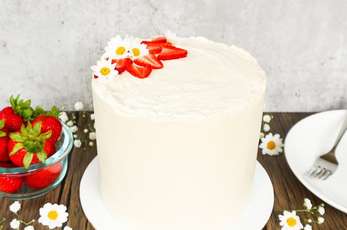 White frosted cake topped with strawberries on a wooden table, with strawberries and a plate beside it