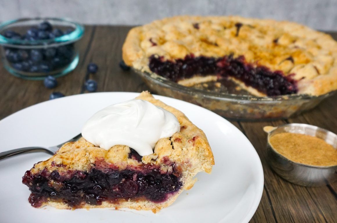 Slice of berry pie with whipped cream on a white plate, beside a whole pie and small bowl on a wooden table