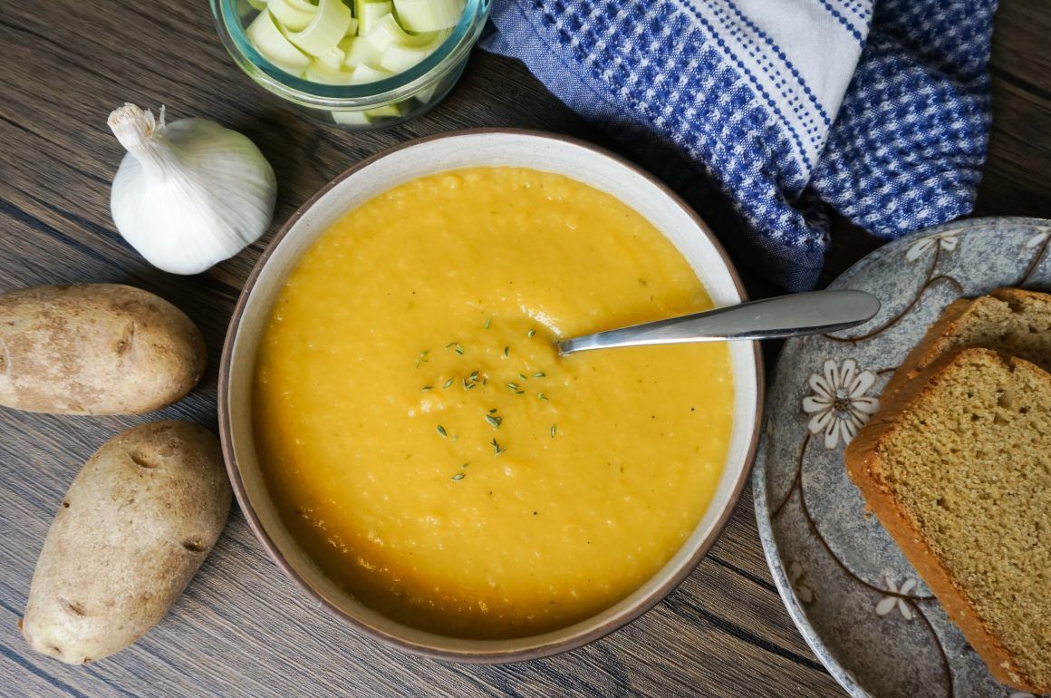 Creamy yellow soup with bread, garlic, potatoes, and a spoon on a rustic table.