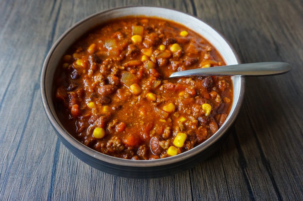Bowl of chunky tomato-based soup or stew with corn, on a wooden table, with a spoon in it