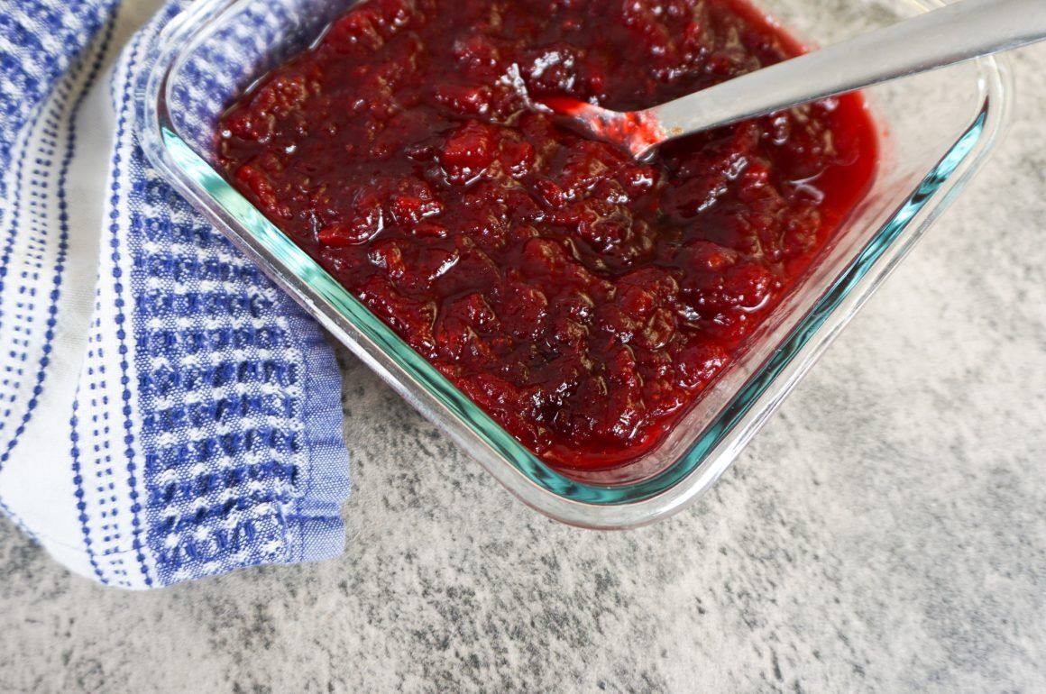 Glass dish of red berry jam with a spoon beside a blue checkered towel on a countertop