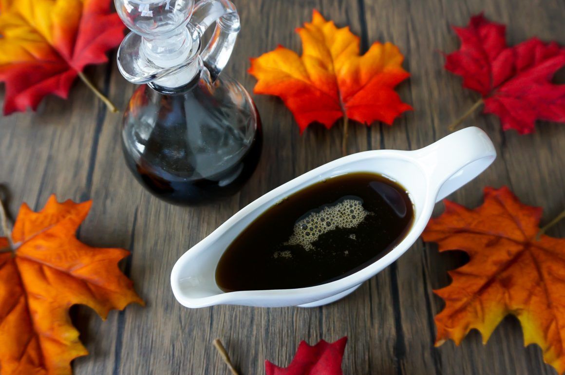 Dark syrup in a white gravy boat on wood, surrounded by colorful autumn leaves and a glass bottle