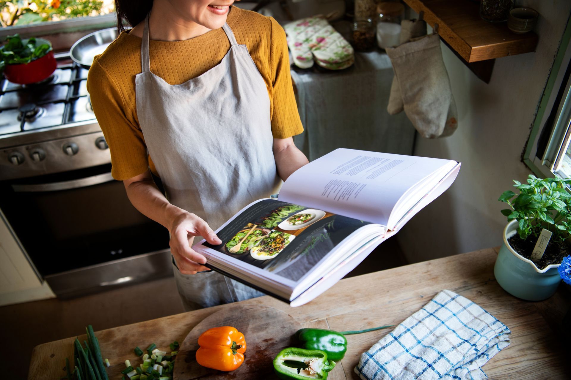 Person in an apron reading a cookbook in a kitchen, with chopped peppers and herbs on the counter.
