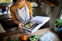 Person in an apron reading a cookbook in a kitchen, with chopped peppers and herbs on the counter.