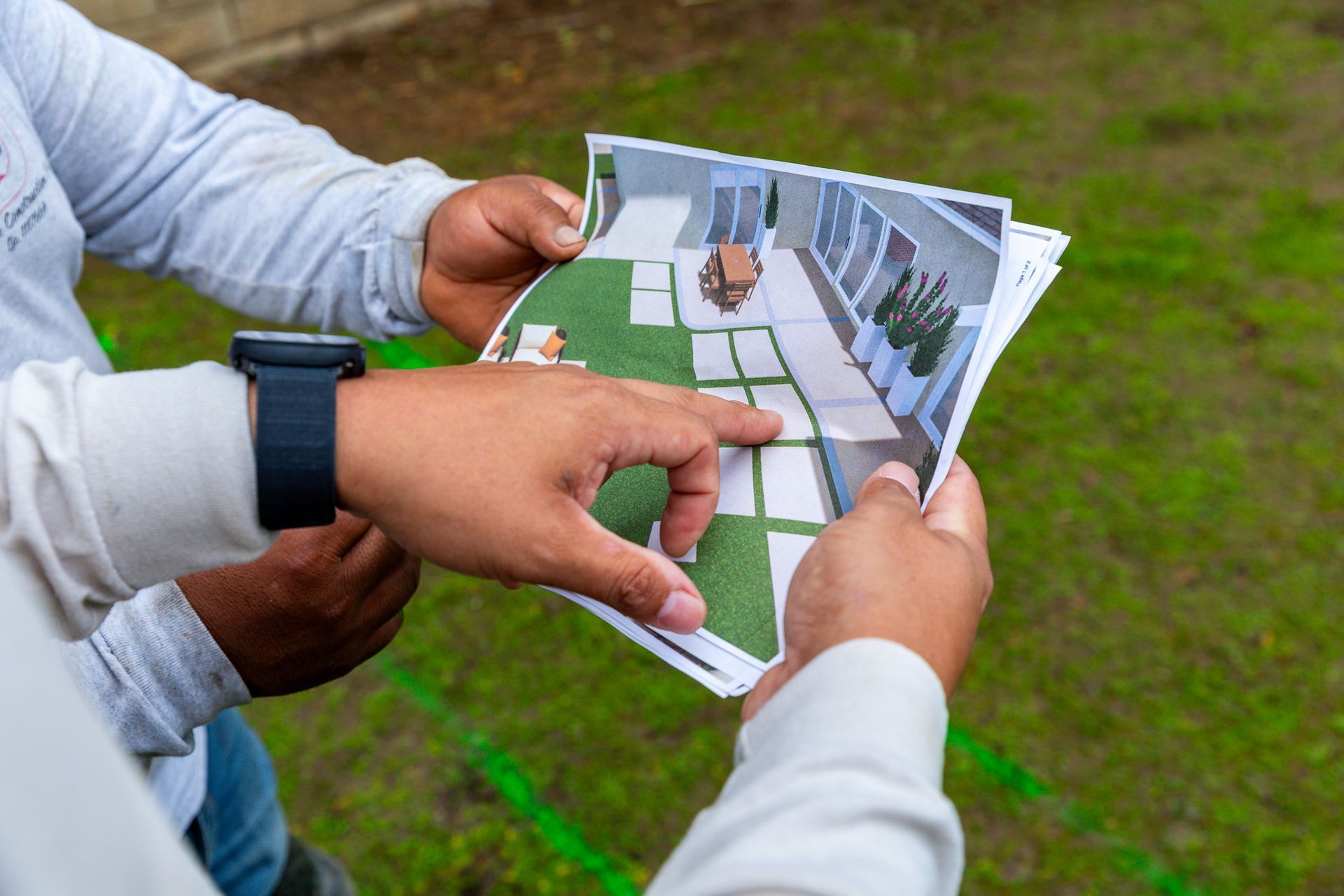 Two people holding a landscaping design print, pointing to a rendered layout of a patio and green space in a backyard.