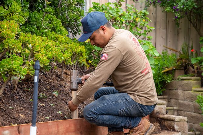 A person in a blue cap and tan long-sleeve shirt uses a mallet to hammer a wooden stake into the ground by a garden bed.