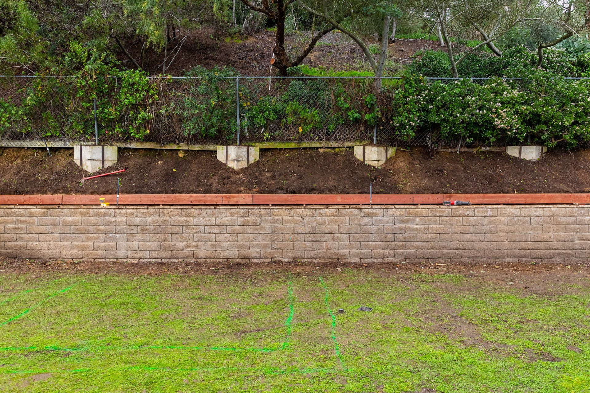 A retaining wall made of tan stone blocks with a wooden header beam, topped by a fence and landscaped hill.