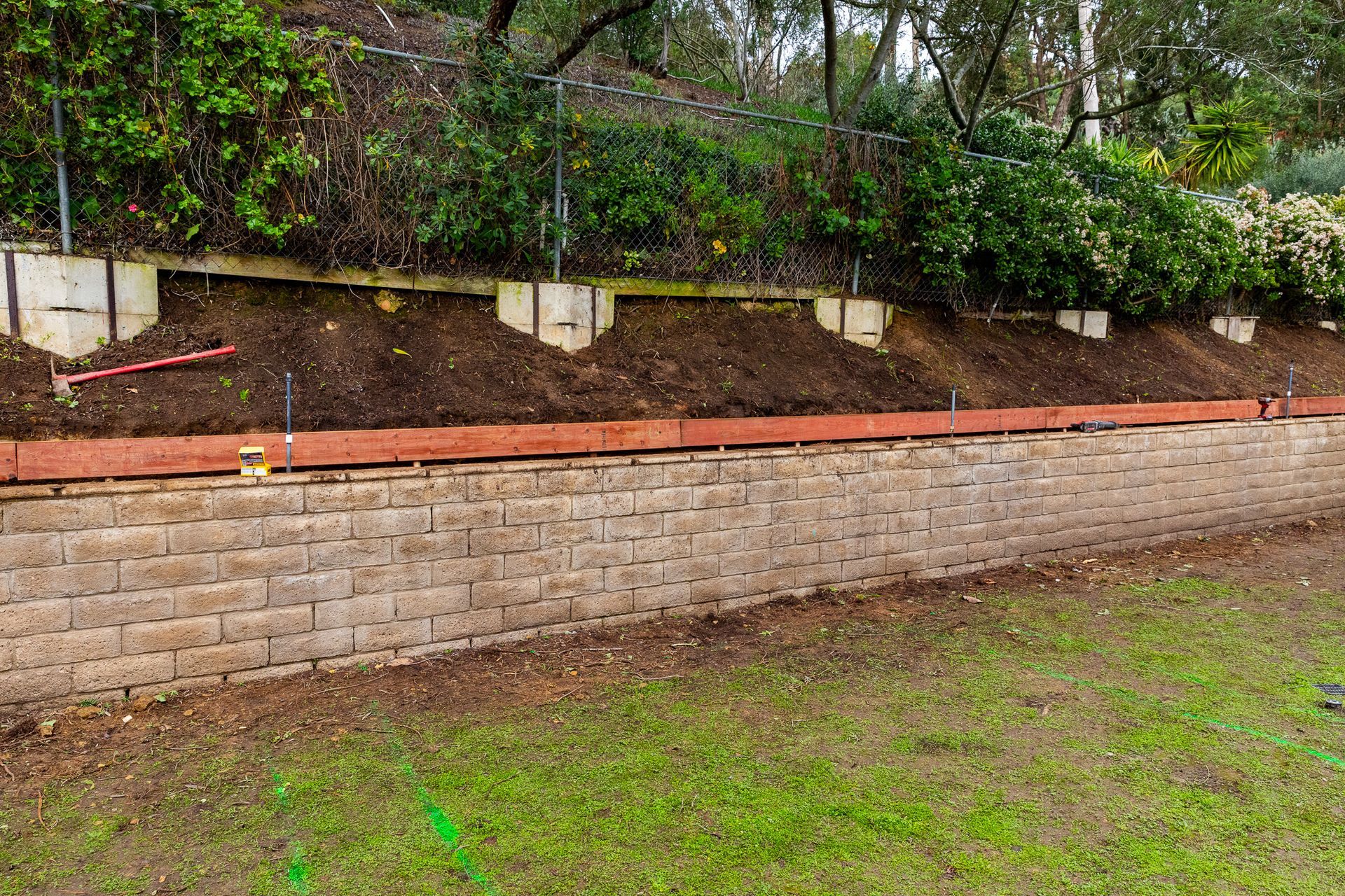 A long, newly constructed tan block retaining wall with a wooden top rail, set against a sloped, terraced garden area.