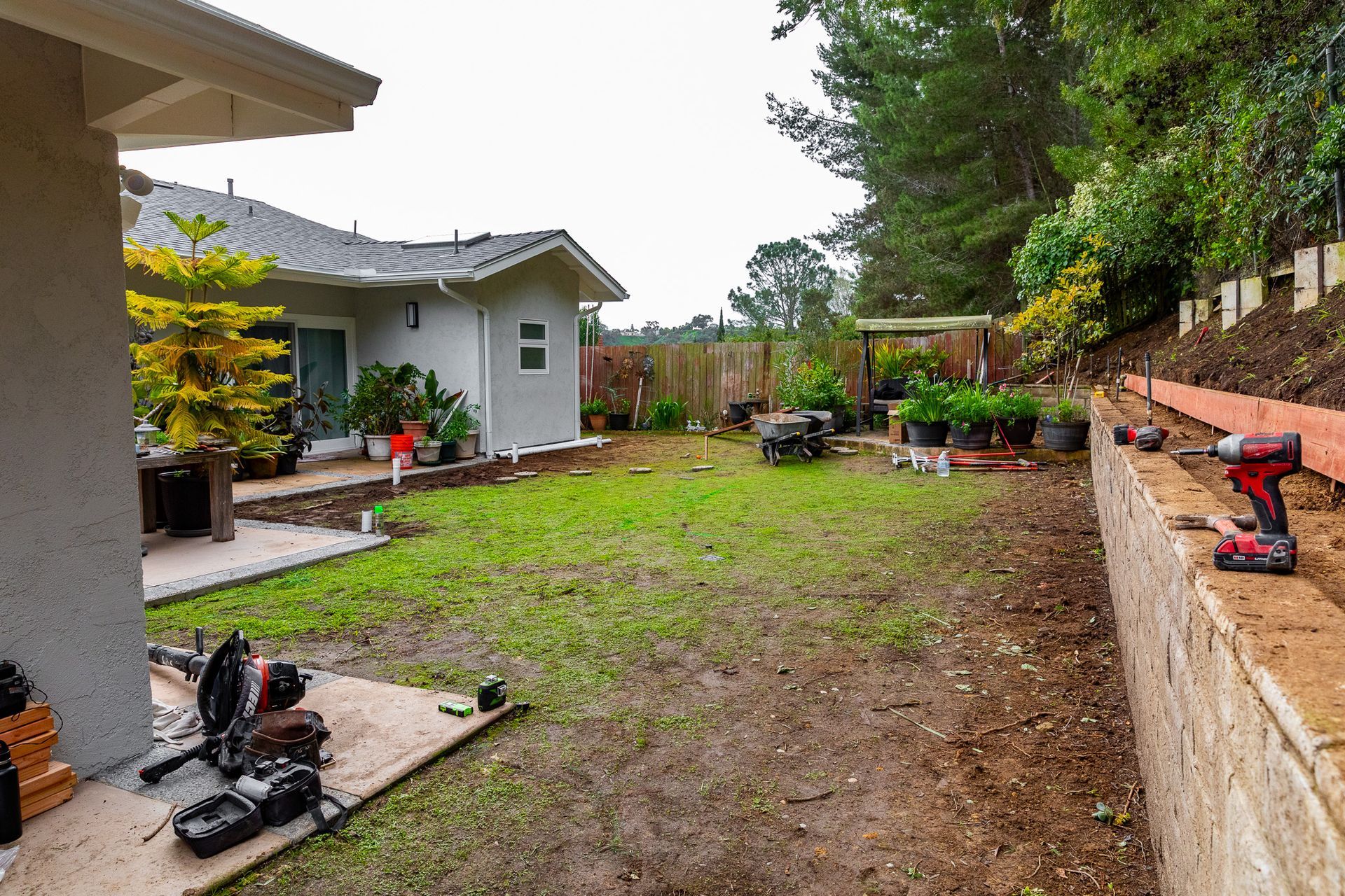 A backyard under construction featuring a concrete retaining wall, patchy lawn, and construction tools on a patio.