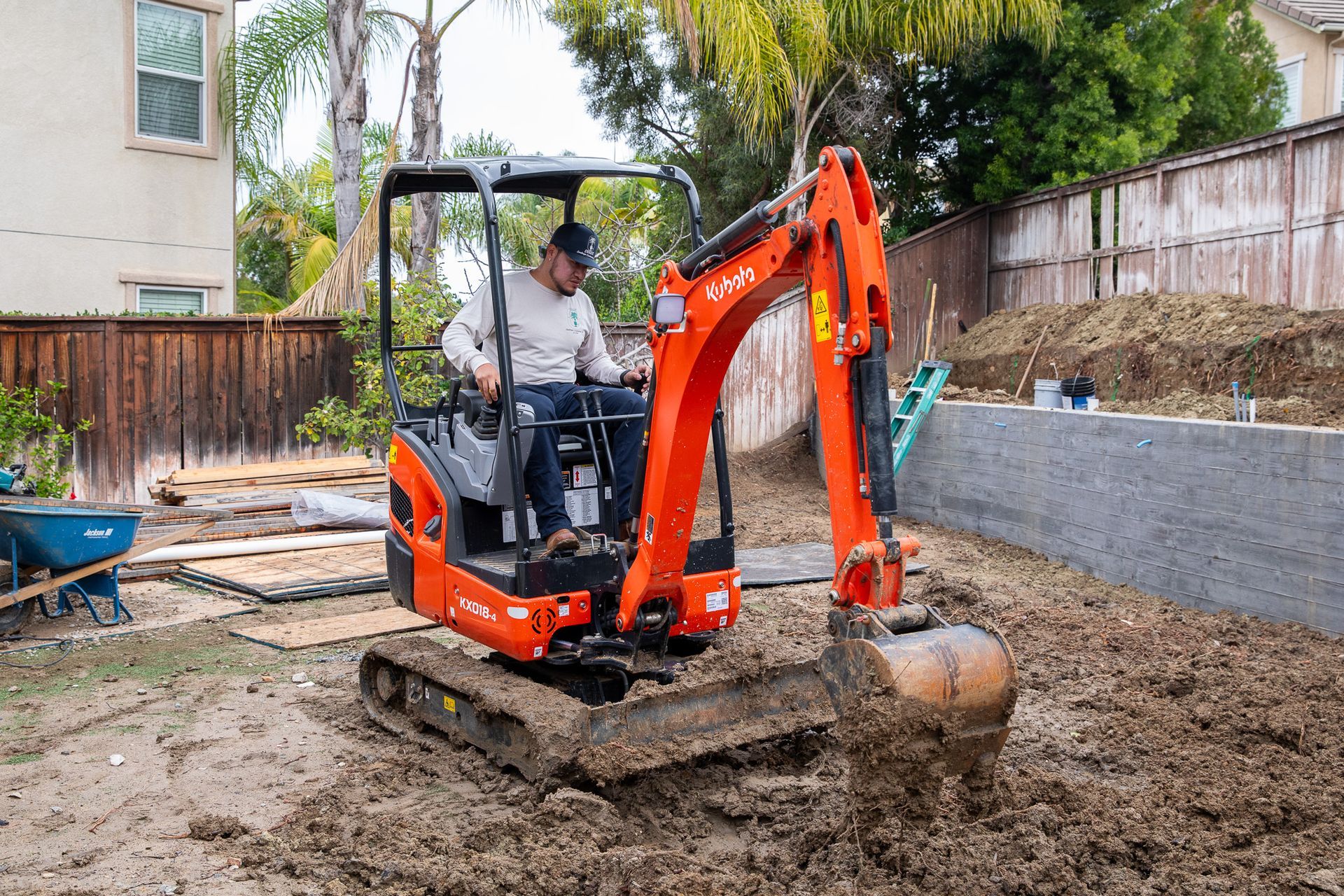 A person operates an orange Kubota mini excavator in a residential backyard with muddy ground.