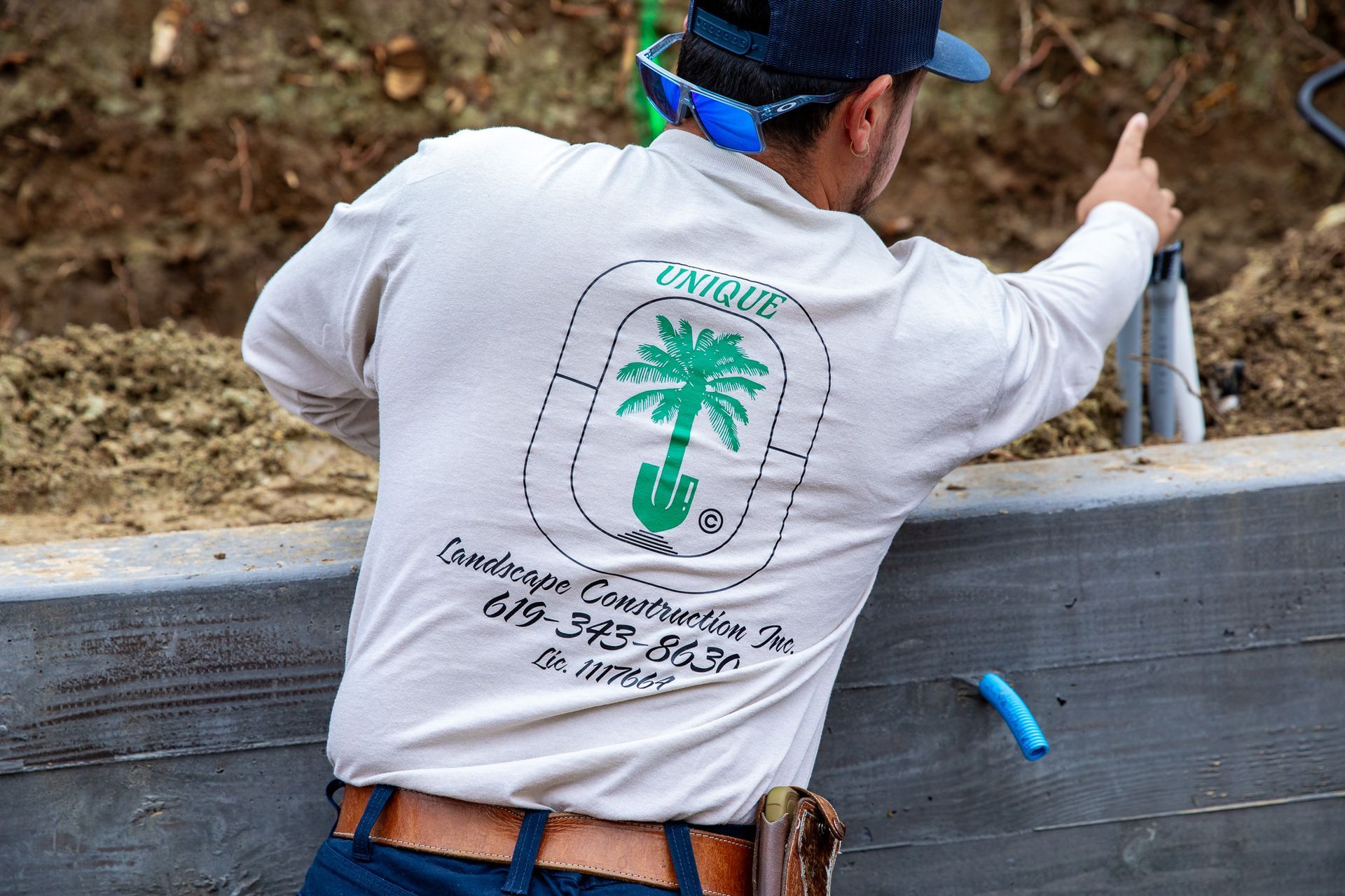 A person in a logo-branded shirt points toward a construction site behind a wooden retaining wall.