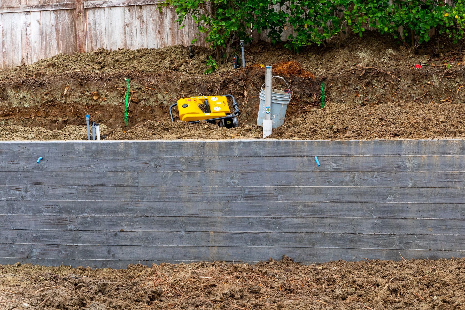 A horizontal, textured concrete retaining wall stands in front of a dirt embankment with a yellow tool and plastic pipes.