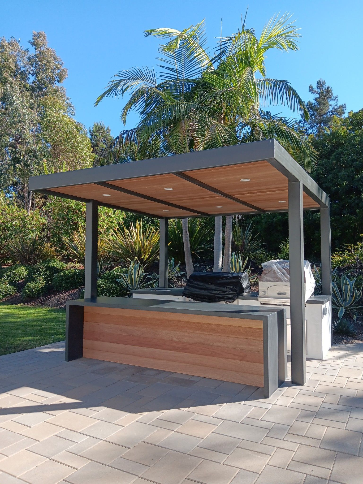 Outdoor kitchen area with a dark metal pergola, wood-paneled island, and grill, set on a stone patio in a lush backyard.